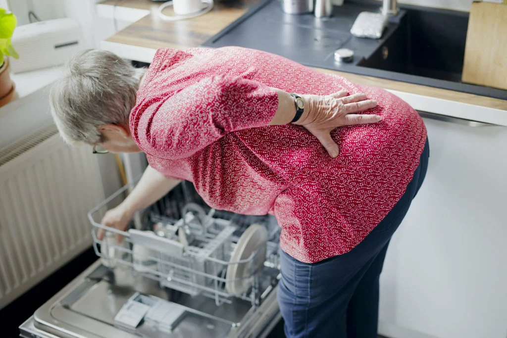 Symbolic photo on the subject of back pain when doing housework. An old woman loads a dishwasher with her back bent, holding her aching back. Berlin, August 31, 2023. (Photo by Thomas Trutschel / Photothek / dpa Picture-Alliance via AFP)